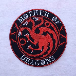 May include: Embroidered patch featuring a red three-headed dragon emblem on a black circular background. The words "MOTHER OF DRAGONS" are embroidered in white around the dragon. Small red dragon silhouettes are also embroidered around the circle.