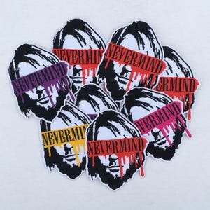 May include: A collection of embroidered patches featuring a stylized face with a black and white design. Each patch has a colored banner across the eyes with the word "NEVERMIND" and dripping paint.