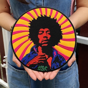 Puede incluir: Parche redondo con un retrato de un músico con un gran afro, en rojo, negro y azul. El fondo tiene rayos amarillos y rosas. El parche tiene un borde negro y texto verde y negro.