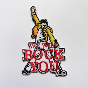 Könnte beinhalten: Ein gestickter Aufnäher mit einer Figur in gelber Jacke und weißer Hose, die einen Arm hebt. Der Text WE WILL ROCK YOU ist in kräftigen roten Buchstaben im unteren Bereich des Designs eingestickt, schwarz umrandet auf weißem Hintergrund.