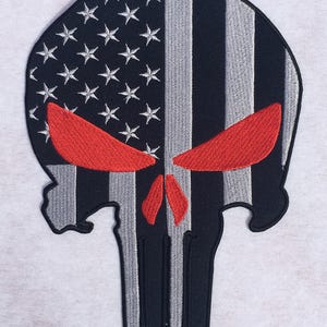 Könnte beinhalten: Ein gestickter Aufnäher in Totenkopfform mit einem schwarz-grauen US-Flaggenmuster, Sternen auf der linken Seite und vertikalen Streifen. Der Totenkopf hat markante, leuchtend rote gestickte Augen und ein rotes Nasendetail.