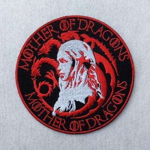 May include: A circular embroidered patch featuring a black background with a red border. The centre displays a white profile of a person with long hair, surrounded by a red dragon silhouette. The text MOTHER OF DRAGONS is stitched in red.
