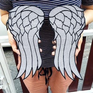 May include: A pair of large, silver-colored embroidered patches shaped like angel wings. Each wing features a detailed, segmented feather design outlined in thick black thread, creating a bold contrast against the metallic silver fabric surface.
