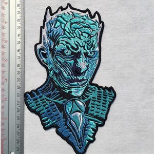 May include: Embroidered patch depicting a character in shades of blue and teal. The patch is approximately 10 cm tall and features detailed facial features and a crown-like design. The design has a textured appearance and is suitable for clothing or accessories.