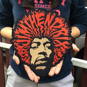 May include: A black patch featuring a portrait of a person with red hair and a tan face. The patch has the word "HEN" embroidered in red. The patch is held up by a person.