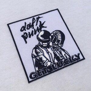 Pode incluir: Um emblema bordado quadrado com fundo branco e borda preta. O design inclui o texto Daft Punk numa fonte estilizada no topo e o texto GET LUCKY na parte inferior, rodeando uma ilustração de duas figuras robóticas.