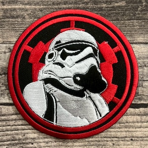 Könnte beinhalten: Runder, gestickter Aufnäher mit einem Stormtrooper-Helm-Design. Der Aufnäher hat einen schwarzen Hintergrund und einen roten Rand. Der Stormtrooper ist in Weiß und Schwarz dargestellt, mit detaillierten Gesichtszügen.