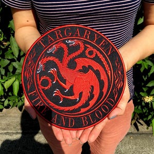 May include: A circular embroidered patch featuring a red three-headed dragon on a black background. The text TARGARYEN is arched across the top, and FIRE AND BLOOD is arched across the bottom in red lettering. The design includes red flame motifs.