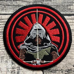 Puede incluir: Parche bordado con un personaje con casco con un diseño de rayos de sol rojos sobre un fondo negro. El parche tiene un borde rojo y un sable de luz blanco. El personaje lleva un atuendo oscuro.