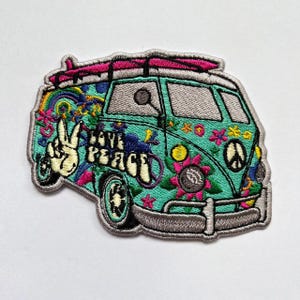 Puede incluir: Un parche bordado con forma de furgoneta color cerceta con una tabla de surf rosa en el techo. El diseño incluye una mano haciendo el signo de la paz, el texto LOVE PEACE, un arcoíris, flores y un símbolo de la paz. Borde gris y costuras detalladas.