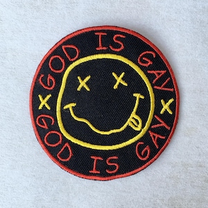 Puede incluir: Parche bordado circular negro con una carita sonriente amarilla, ojos tachados y lengua fuera. Las palabras "GOD IS GAY" están bordadas en rojo alrededor de la carita sonriente. El parche tiene un borde rojo.