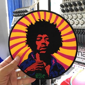 Puede incluir: Un parche bordado circular que presenta el retrato estilizado de una persona con un gran peinado afro. El fondo consiste en rayas radiantes de color amarillo y magenta. La persona viste una camisa azul con un patrón estampado.