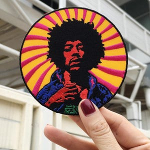 Może przedstawiać: Okrągła, haftowana naszywka przedstawiająca portret muzyka z dużą afro. Naszywka ma żółto-różowy wzór promieni słonecznych wokół portretu. Muzyk ma na sobie niebieską kurtkę. Naszywka jest trzymana w dłoni z czerwonym lakierem do paznokci.
