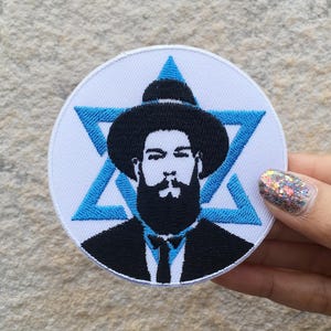 May include: A circular embroidered patch featuring a white background with a bright blue Star of David. Centered on the patch is a black and white portrait of a bearded individual wearing a wide-brimmed hat, a suit jacket, and a necktie.