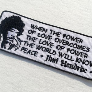 Op de afbeelding: Geborduurde patch met een zwart-wit afbeelding van Jimi Hendrix en de quote: "When the power of love overcomes the love of power, the world will know peace." De patch heeft een witte achtergrond.