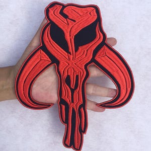 Puede incluir: Un parche bordado en rojo y negro que presenta el cráneo estilizado de un mythosaur. El diseño consiste en un cráneo central con dos cuernos curvos que se extienden hacia afuera y una mandíbula inferior puntiaguda, todo delineado en hilo negro.