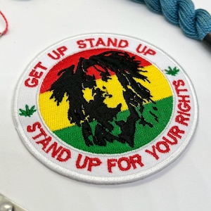 May include: Embroidered patch with a circular design featuring the text "GET UP STAND UP STAND UP FOR YOUR RIGHTS" in red and white. The patch has a black silhouette over a background of red, yellow, and green, with green leaf accents.