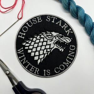 May include: A black and white embroidered patch featuring the House Stark direwolf sigil from Game of Thrones. The text "HOUSE STARK" and "WINTER IS COMING" are embroidered around the wolf's head. A needle, thread, and scissors are in the image.