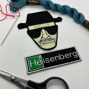 May include: Embroidered patches featuring a character with a black hat and sunglasses, and the name "Heisenberg". The patches are black, white, and beige. A spool of blue thread, a needle, and scissors are also visible.