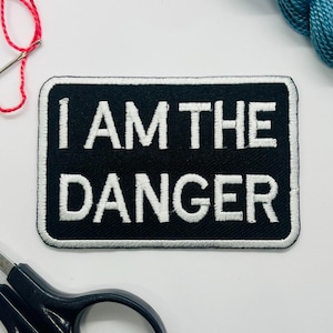 May include: Black rectangular patch with white embroidered text that reads "I AM THE DANGER." The patch has a white border. A needle with red thread, scissors, and blue thread are in the background.