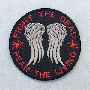 May include: A circular black embroidered patch featuring two light gray angel wings in the center. The red text "FIGHT THE DEAD" is arched across the top, and "FEAR THE LIVING" is arched across the bottom, with small red cross symbols.