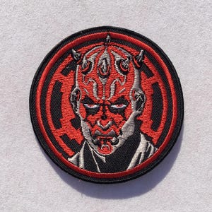 May include: A circular embroidered patch featuring a detailed portrait of a character with red and black facial markings, horns, and intense eyes. The design is set against a red and black circular background with a stylized emblem pattern.
