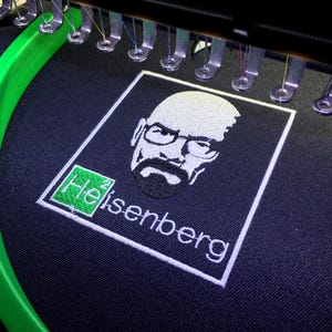 Op de afbeelding: Geborduurde patch met een gestileerd portret van een man met een bril en een sik, met de naam "Heisenberg" eronder. Het ontwerp is in witte draad op een donkerblauwe stof, met een groen en wit element in de hoek. De patch bevindt zich in een borduurmachine.