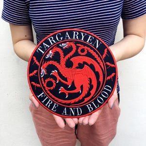 Könnte beinhalten: Ein runder, schwarzer Aufnäher mit den weißen Worten "TARGARYEN FIRE AND BLOOD". Der Aufnäher zeigt ein rotes, dreiköpfiges Drachenemblem. Kleine rote fledermausartige Figuren umgeben den Drachen.