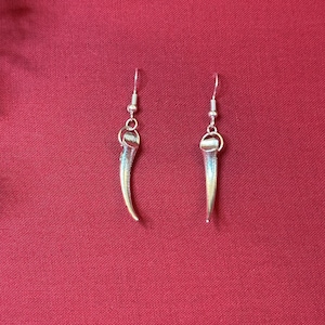 May include: A pair of silver-colored earrings, each featuring a curved, claw-like design. The earrings hang from a simple hook. The background is a solid, deep red fabric.