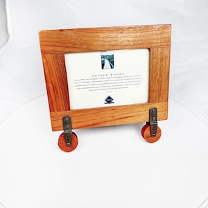 May include: A rectangular wooden picture frame with a natural grain finish. The frame features two metal brackets attached to the bottom, each holding a small wooden wheel. A white insert card with the text SHAKER WOODS is centered in the frame.