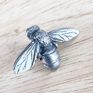 Puede incluir: Figurita detallada de abeja plateada con alas y cuerpo texturizados. La abeja está colocada sobre una superficie de madera clara. Las alas están extendidas y la abeja parece estar en pleno vuelo. La figura es un objeto decorativo.
