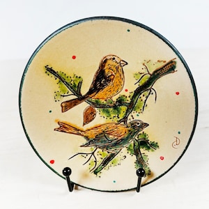 May include: Round ceramic plate with a hand-painted design featuring two birds perched on a branch. The birds are depicted in shades of brown and yellow, with green foliage and red and green accents. The plate has a cream-colored background and a dark green rim.