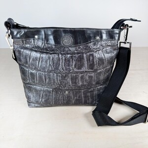 May include: A black and gray crossbody bag with a textured, crocodile-like pattern. The bag has a black adjustable strap, silver-tone hardware, and a decorative emblem on the front. The bag is made of leather.