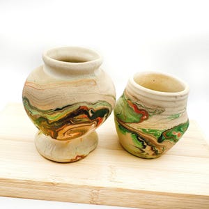 May include: Two ceramic vases with a marbled design in shades of beige, green, orange, and brown. One vase is round with a pedestal base, and the other is a smaller, tilted cylinder. Both are on a light wood surface.