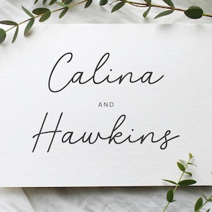 Puede incluir: Una tarjeta rectangular blanca con los nombres "Calina" y "Hawkins" en elegante escritura negra. La palabra "AND" está centrada entre los nombres. La vegetación, incluyendo ramitas y hojas, enmarca la tarjeta, creando una estética natural.