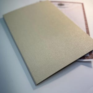 May include: A light beige, textured book cover with a rectangular shape. The cover is slightly angled, revealing a glimpse of a document with a decorative border underneath. The book cover is the primary focus.