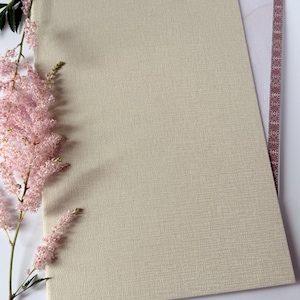 Può includere: Un cartoncino rettangolare color crema con superficie testurizzata, che si sovrappone parzialmente a un documento con bordo a motivi marroni e il testo "LATVIJAS". Un rametto di fiori di astilbe rosa chiaro con steli verdi è posizionato a sinistra.