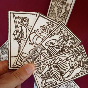 Może przedstawiać: Zestaw prostokątnych kart z ilustracjami wykonanymi czarnym tuszem na białym tle. Karty przedstawiają różne postacie w historycznych strojach, w tym osobę trzymającą miecz, osobę z pentaklem oraz ozdobne wzory dekoracyjne.