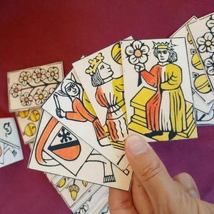 Peut inclure: Un ensemble de cartes à jouer rectangulaires ornées de dessins au trait à l'encre noire avec des touches d'aquarelle jaune et orange. Les cartes présentent diverses illustrations dont un personnage couronné tenant une fleur, un écu et des motifs floraux.