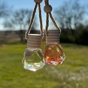 Puede incluir: Dos ambientadores colgantes para coche con frascos de vidrio transparentes y llenos de purpurina. Los frascos en forma de diamante est&aacute;n suspendidos por una cuerda con cuentas de madera. Un frasco es transparente, el otro est&aacute; lleno de purpurina naranja.