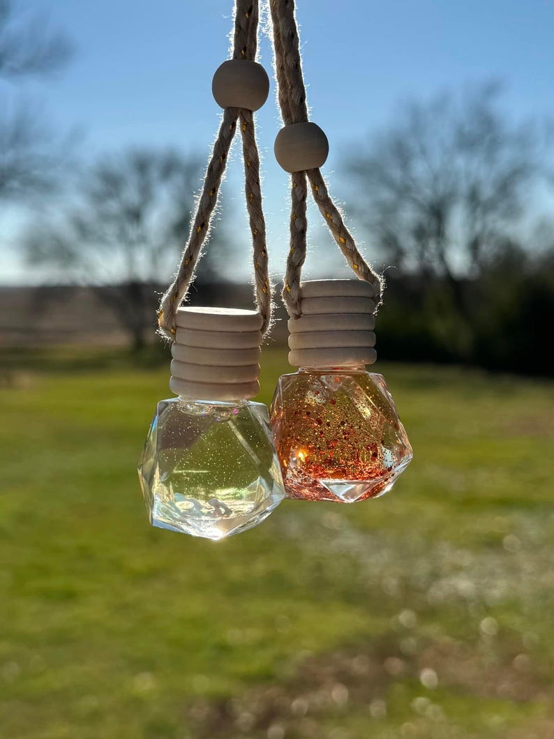 Puede incluir: Dos ambientadores colgantes para coche con frascos de vidrio transparentes y llenos de purpurina. Los frascos en forma de diamante est&aacute;n suspendidos por una cuerda con cuentas de madera. Un frasco es transparente, el otro est&aacute; lleno de purpurina naranja.