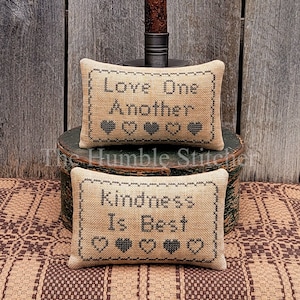 May include: Two small, rectangular, beige pillows with black embroidery. The top pillow reads "Love One Another" with black hearts. The bottom pillow reads "Kindness Is Best" with black hearts. The pillows are sitting on a wooden box with a candle holder on top.