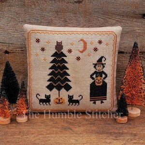 May include: A decorative Halloween-themed pillow with a cross-stitch design featuring a witch, black cats, and a tree. The pillow is surrounded by small decorative trees in black and orange. The text "The Humble Stitcher" is at the bottom.
