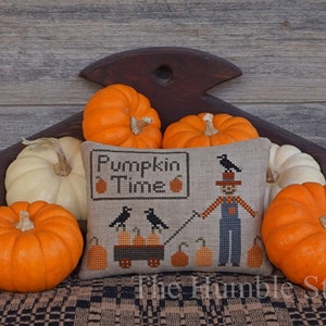 Pode incluir: Uma almofada de ponto cruz com o texto "Pumpkin Time" e um design de espantalho. A almofada é cercada por abóboras laranja e brancas.