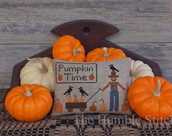 May include: A cross-stitch pillow with the text "Pumpkin Time" and a scarecrow design. The pillow is surrounded by orange and white pumpkins.