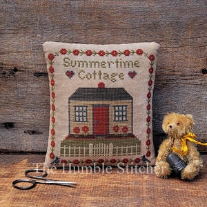 May include: A square beige fabric pincushion featuring a cross-stitched design of a house with a red door, a white fence, and red flowers. The text reads Summertime Cottage. A small brown teddy bear with a yellow ribbon and a metal thimble sits nearby.