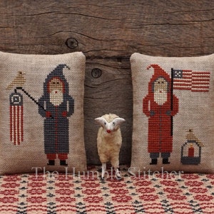 May include: Two embroidered pillow covers with a rustic, vintage aesthetic. One features a Santa Claus holding a flag with the text "The Humble Stitcher". The other features a Santa Claus holding a lantern.