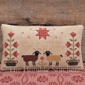 Pode incluir: Uma almofada retangular de tecido bege com um design em ponto cruz de duas ovelhas, uma castanha e uma bege, entre duas flores vermelhas em vasos. Uma estrela vermelha está acima das ovelhas. O texto The Humble Stitcher está na parte inferior.