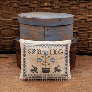 May include: A beige pillow with a cross-stitch design featuring a flower pot with a plant and the word "SPRING" above it. The pillow is sitting on top of a blue wooden box.