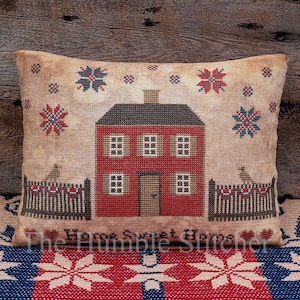 May include: A cross-stitch pillow featuring a red house with white windows and a black door. The house is surrounded by a black picket fence with a red, white, and blue bunting. The background is a tan fabric with red and blue stars. The text "Home Sweet Home" is stitched below the house.